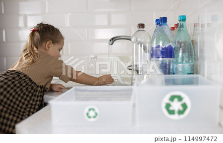 Against the background of boxes with a recycling sign, a child sorts plastic. A small child helps put plastic bottles into boxes for recycling. Concept of sorting plastic for recycling. Against the background of boxes with a recycling sign, a child sorts plastic. A small child helps put plastic bottles into boxes for recycling. Concept of sorting plastic for recycling. 114997472