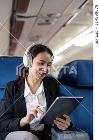 Businesswoman using tablet and headphones on airplane. Concept of in flight productivity and technology Businesswoman using tablet and headphones on airplane. Concept of in flight productivity and technology 114998678