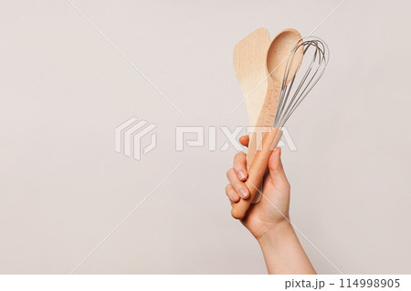 Hand holding a wooden spoon, a wooden spatula, and a whisk against a plain beige background, copy space 114998905