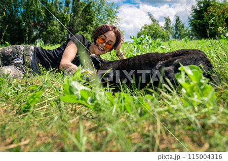 Beautiful happy girl lies and plays on the lawn with her dog. Young woman cuddling her dog on the grass. 115004316