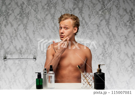 Young shirtless man standing in bathroom and looking at mirror with contemplative expression against marbled wall. 115007081