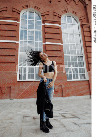 A woman in a black jacket and jeans poses in front of a brick building. Fashon street shot. A woman in a black jacket and jeans poses in front of a brick building. Fashon street shot. 115009893