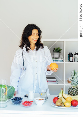 Confident smiling middle aged female Indian Nutritionist doctor holding orange in front of office desk full of fruits and vegetables. Healthy diet habits and wellness lifestyle, weight loss program. Confident smiling middle aged female Indian Nutritionist doctor holding orange in front of office desk full of fruits and vegetables. Healthy diet habits and wellness lifestyle, weight loss program. 115010235
