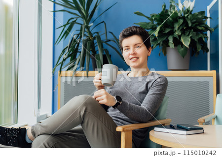 Smiling Neutral gender middle aged business woman in casual clothing relaxing with coffee cup on the armchair. Female professional enjoying coffee drink in office lounge. Take a break for recovery 115010242