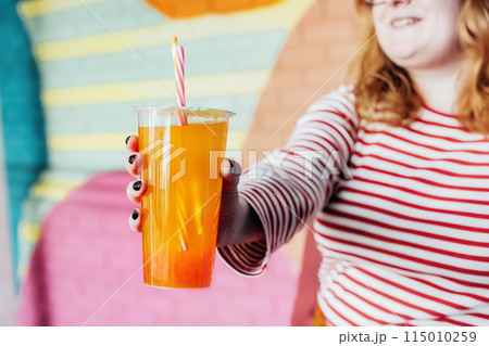 Glass of fruity sugar flavored tapioca pearl orange bubble tea with straw in hand of hipster fashion woman in bright clothes on the colorful painted brick wall background. Selective focus. Copy space. Glass of fruity sugar flavored tapioca pearl orange bubble tea with straw in hand of hipster fashion woman in bright clothes on the colorful painted brick wall background. Selective focus. Copy space. 115010259