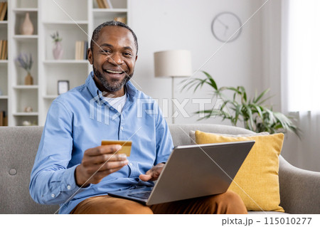 Smiling man holding a credit card, using laptop for online shopping at home. Comfortable living room setting with shelves and plant background. Lifestyle, technology, finance. Smiling man holding a credit card, using laptop for online shopping at home. Comfortable living room setting with shelves and plant background. Lifestyle, technology, finance. 115010277