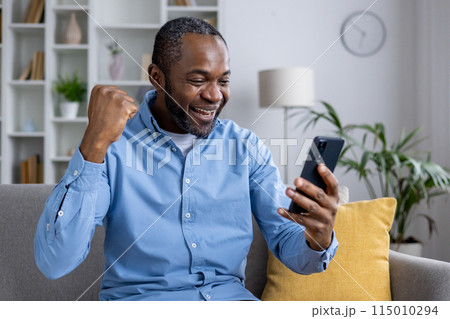 Joyful African American man in a blue shirt cheering with a clenched fist while looking at his smartphone in a cozy living room. 115010294