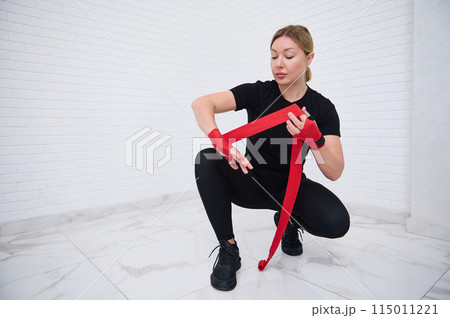 Authentic portrait of determined confident European young woman fighter 40s, in sports clothes, sitting in squat position, typing red tapes over white background, preparing for boxing training Authentic portrait of determined confident European young woman fighter 40s, in sports clothes, sitting in squat position, typing red tapes over white background, preparing for boxing training 115011221