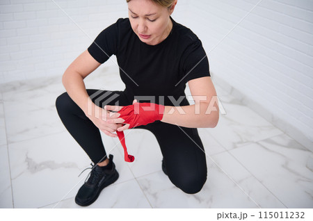 Determined confident European boxer fighter woman 40s, in black sports wear, tying tape around her hand before get boxing gloves preparing to boxing practice. Martial art and combat concept 115011232