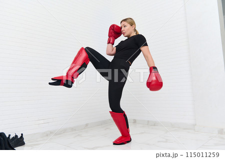 European blonde young kickboxing woman in black activewear and red kickboxing gloves, isolated white background performing martial arts kick. Sport exercise, fitness and workout. Full length portrait European blonde young kickboxing woman in black activewear and red kickboxing gloves, isolated white background performing martial arts kick. Sport exercise, fitness and workout. Full length portrait 115011259