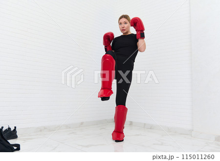 European blonde young kickboxing woman in black activewear and red kickboxing gloves, isolated on white background performing a martial arts kick. Sport exercise, fitness and cardio workout workout. European blonde young kickboxing woman in black activewear and red kickboxing gloves, isolated on white background performing a martial arts kick. Sport exercise, fitness and cardio workout workout. 115011260
