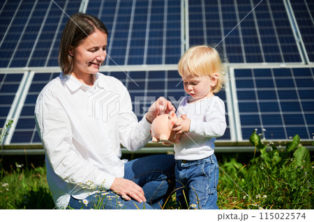 Modern mom and her little kid saving money in piggy bank. Mother teaching son how to invest money in future. Smiling mom and her kid putting savings into piggybank on background of solar panels. Modern mom and her little kid saving money in piggy bank. Mother teaching son how to invest money in future. Smiling mom and her kid putting savings into piggybank on background of solar panels. 115022574