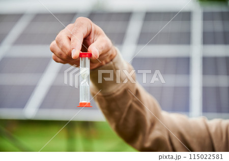 Time working for green energy investments. Solar batteries generating energy that generating profit. Sand clock in man hand on background of solar PV battery. Close up Time working for green energy investments. Solar batteries generating energy that generating profit. Sand clock in man hand on background of solar PV battery. Close up 115022581