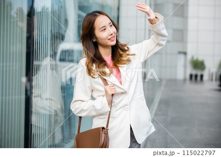 Portrait of confidence young businesswoman standing outside office building in city raise your hand to shade the sun. Happy woman wearing white suit jacket with brown bag at sunlight outdoors. 115022797