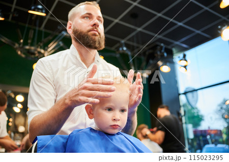 Young child getting haircut in modern barbershop. Hairdresser making final touches in new haircut, shaping child's hair by hands. Young child getting haircut in modern barbershop. Hairdresser making final touches in new haircut, shaping child's hair by hands. 115023295