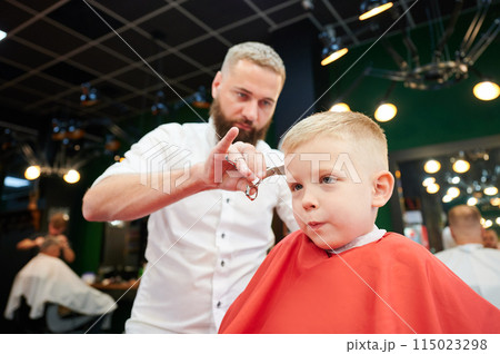 Man barber cutting little boy's hair using comb and scissors. Child getting haircut from adult male, likely barber. Professional hairdresser and cute client at modern barbershop. 115023298