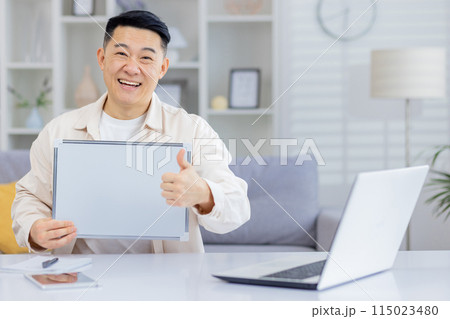 A cheerful man giving a thumbs up while holding a blank board at a home office setup with a laptop, portraying positivity and success. 115023480
