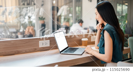 A woman is sitting at a table with a laptop in front of her. She is looking at the screen and she is focused on her work. The scene takes place in a cafe, with other people visible in the background A woman is sitting at a table with a laptop in front of her. She is looking at the screen and she is focused on her work. The scene takes place in a cafe, with other people visible in the background 115025690