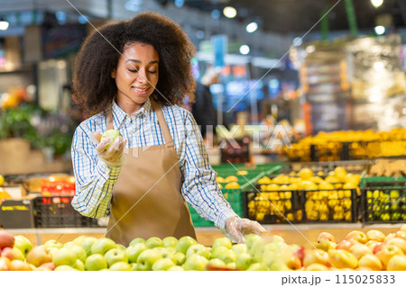 Smiling woman in apron sorting fresh apples at a grocery store, showcasing a vibrant produce section 115025833