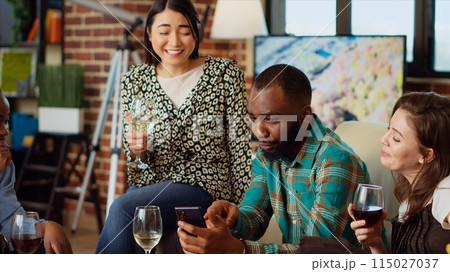 Multiracial friends at apartment party looking at funny videos on cellphone screen, sharing their impressions. Joyful guests in living room reacting to online clips on mobile phone Multiracial friends at apartment party looking at funny videos on cellphone screen, sharing their impressions. Joyful guests in living room reacting to online clips on mobile phone 115027037