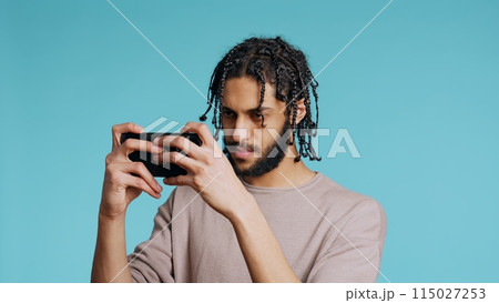 Gamer disappointed after receiving game over screen on cellphone touchscreen display. Middle Eastern man nodding head, upset after losing videogame, playing on phone, studio background, camera B 115027253