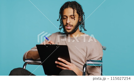 Middle Eastern man in wheelchair suffering from paraplegia holding clipboard in hand, using pen to write on paper. Person with disability completing forms, isolated over studio background, camera B Middle Eastern man in wheelchair suffering from paraplegia holding clipboard in hand, using pen to write on paper. Person with disability completing forms, isolated over studio background, camera B 115027254
