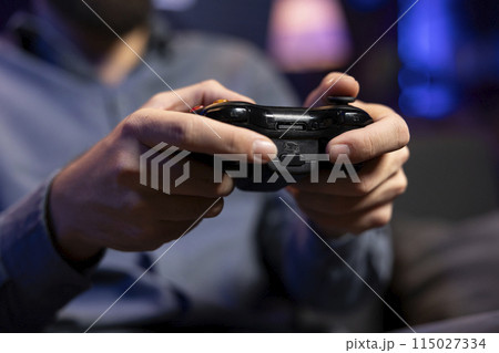 Focus shot on controller held by gamer in blurry background playing videogames. Close up shot of gamepad used by man enjoying gaming session in living room while sitting down in front of TV 115027334