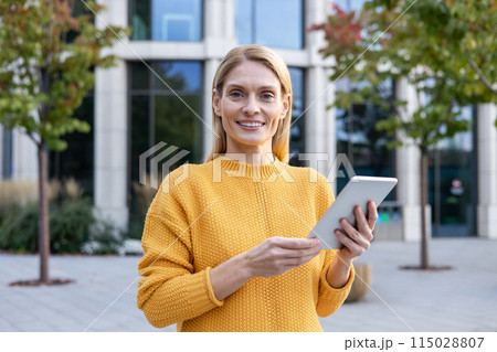 Confident professional woman in a yellow sweater using a tablet outside a modern office building. Represents technology in everyday business. 115028807