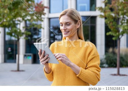 A middle-aged professional woman uses a digital tablet outside a modern building. Dressed in a vibrant yellow sweater, she appears focused and content. 115028813
