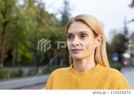 Close-up of a mature Caucasian woman in a yellow sweater, looking contemplative as she enjoys a peaceful autumn day outdoors. The background is softly blurred with hints of greenery. Close-up of a mature Caucasian woman in a yellow sweater, looking contemplative as she enjoys a peaceful autumn day outdoors. The background is softly blurred with hints of greenery. 115028872