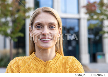 A portrait of a confident middle-aged businesswoman smiling outdoors. She is wearing a yellow sweater, with a modern office building in the background. 115028874