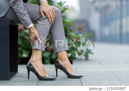 A close-up image of a businesswoman touching her calf, showing discomfort or pain in her leg, seated outdoors in an urban setting. 115028897