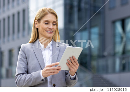 A successful professional businesswoman in a gray suit smiles while using a tablet in a modern urban setting, representing empowerment in the corporate world. She exudes confidence and competence A successful professional businesswoman in a gray suit smiles while using a tablet in a modern urban setting, representing empowerment in the corporate world. She exudes confidence and competence 115028916