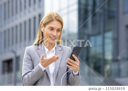 A confident businesswoman in a grey suit talks on her smartphone with earphones outside a modern office building. She showcases a professional and focused demeanor, embodying success in her career A confident businesswoman in a grey suit talks on her smartphone with earphones outside a modern office building. She showcases a professional and focused demeanor, embodying success in her career 115028919