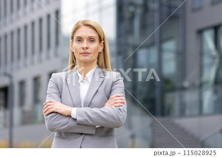 Confident businesswoman in a grey suit standing with her arms crossed outside a modern office building. Confident businesswoman in a grey suit standing with her arms crossed outside a modern office building. 115028925