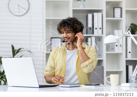 Smiling man wearing a headset while working on a laptop in a modern office setting. Professional workspace with office supplies and plants. Smiling man wearing a headset while working on a laptop in a modern office setting. Professional workspace with office supplies and plants. 115037213