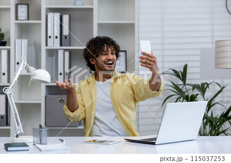 Young man in a modern office taking a selfie with his smartphone, expressing joy and confidence while working at his desk with a laptop. 115037253