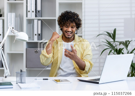 Excited young man celebrating success while looking at smartphone in a modern office setting. Achieving goals and experiencing happiness at work. 115037266