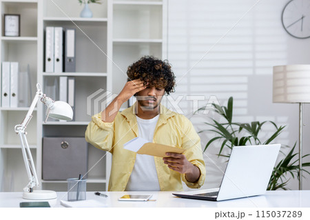 Young man reading mail envelope and receiving bad news in a home office setting, feeling stressed and worried. Modern workspace with laptop and office supplies. Young man reading mail envelope and receiving bad news in a home office setting, feeling stressed and worried. Modern workspace with laptop and office supplies. 115037289