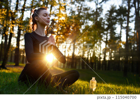 A young beautiful woman in a tight-fitting tracksuit sits on the grass in the woods in lotus pose and meditates. A young beautiful woman in a tight-fitting tracksuit sits on the grass in the woods in lotus pose and meditates. 115040700