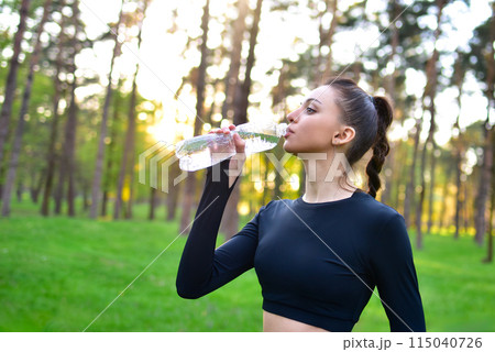 A woman in a sporty black top holds up a water bottle for a drink of water after a sport in the park. 115040726