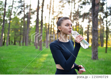 A woman in a sporty black top holds up a water bottle for a drink of water after a sport in the park. 115040729