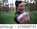 sporty woman doing martial arts training in sporty black top holding hands in fighting pose against forest background. 115040733