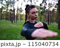 A young woman in a black training clothing holds her arms in a fighting pose for battle in the forest. 115040734