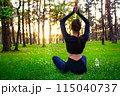 Slim beautiful woman in black tight fitting tracksuit sitting on grass in forest in yoga pose. back view 115040737