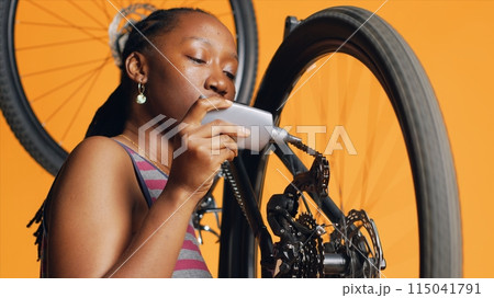 BIPOC mechanic using specialized glue to fix damaged bicycle chain, orange studio background. Expert applying adhesive on bike parts during checkup process, close up shot 115041791