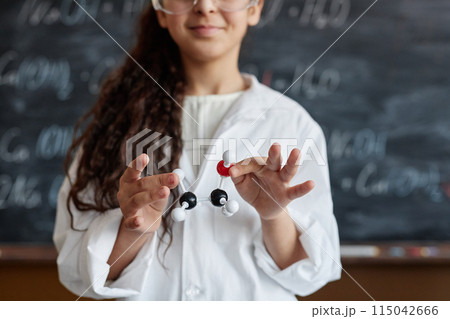Medium closeup of schoolgirl wearing lab coat standing against blackboard holding chemical structure model in hands 115042666