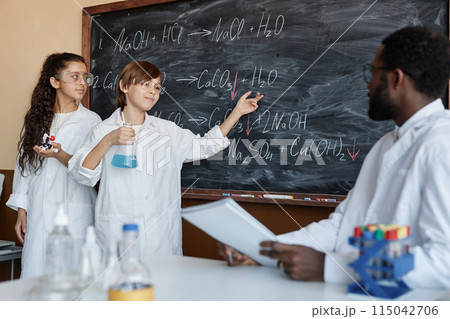 Two diverse students wearing laboratory coats standing at blackboard in classroom solving problem in Chemistry class Two diverse students wearing laboratory coats standing at blackboard in classroom solving problem in Chemistry class 115042706