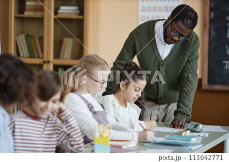 Young African American teacher standing at desk in classroom checking on girl doing grammar exercise during lesson Young African American teacher standing at desk in classroom checking on girl doing grammar exercise during lesson 115042771