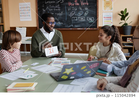 Young African American teacher of English sitting at table in classroom showing alphabet cards to kids during lesson Young African American teacher of English sitting at table in classroom showing alphabet cards to kids during lesson 115042894